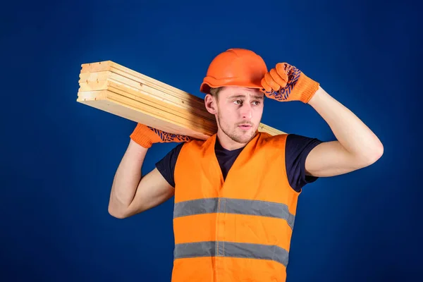 Menuisier, menuisier, ouvrier, constructeur porte une poutre en bois sur l'épaule. L'homme en gants de protection tient la visière du casque, corrige le casque dur sur la tête, fond bleu. Concept de menuisier . — Photo