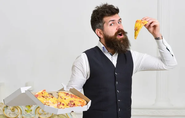 Macho in classic clothes hungry, on cheerful face, holds slice of pizza, eating, white background. Pizza delivery concept. Man with beard and mustache holds delivered box with tasty fresh hot pizza.