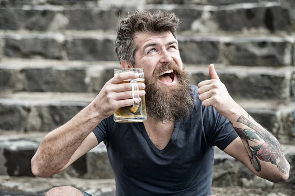 Homem barbudo segura caneca de cerveja, bebe cerveja ao ar livre. Conceito de cerveja artesanal. Homem com barba longa parece relaxado, mostra o polegar para cima. Homem com barba e bigode no rosto feliz, fundo pedregoso, desfocado . — Fotografia de Stock