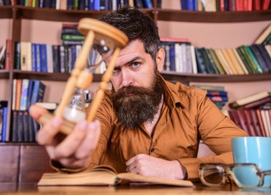 Düşünceli yüzlü bir adam ders çalışırken kum saati, arka planda kitaplık tutuyor. Kendi kendine eğitim kavramı. Adam bardak ve bardaklarla masada oturuyor. Öğretmen ya da sakallı öğrenci kütüphanede çalışıyor..