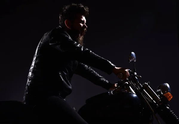 Man with beard, biker in leather jacket sitting on motor bike in darkness, black background. Macho, brutal biker in leather jacket riding motorcycle at night time, copy space. Masculinity concept. — Stock Photo, Image