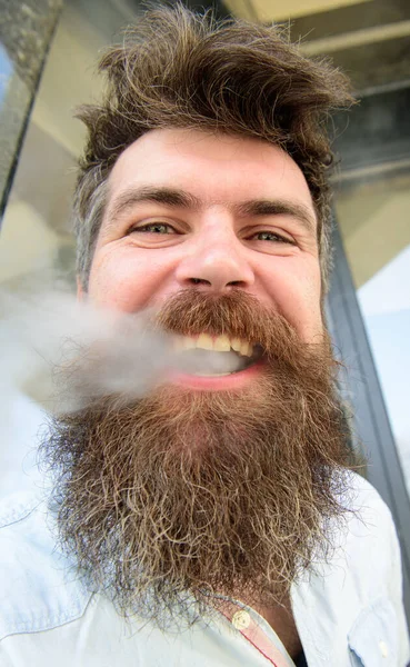 Hipster with tousled hair and gray on relaxed cheerful face with white smoke flying out of mouth. Man with beard and mustache smoking, building background. Vaping concept. — Stock Photo, Image