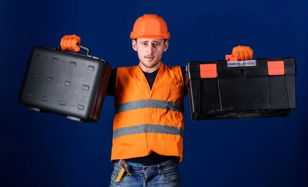 Caixa de ferramentas e conceito de equipamento. Homem de capacete, chapéu duro segura caixa de ferramentas e mala com ferramentas, fundo azul. Trabalhador, reparador, reparador, construtor no equipamento pensativo da escolha da cara para o trabalho . — Fotografia de Stock