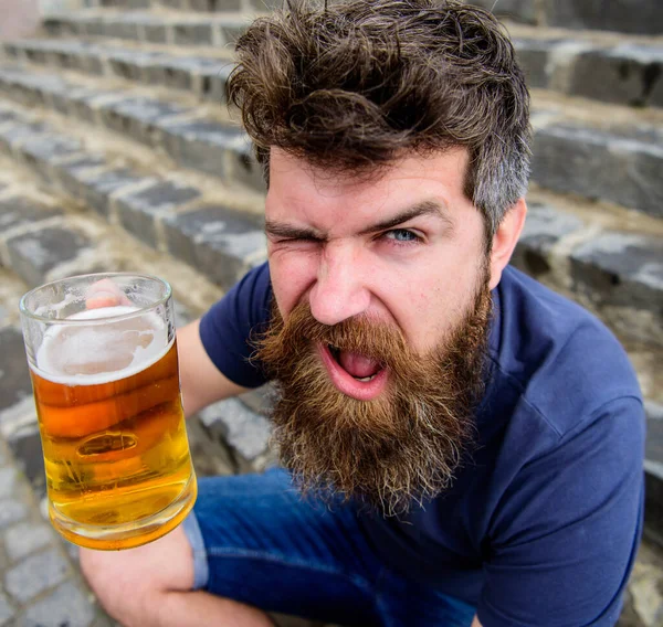 Ünnepség koncepció. Egy fickó, aki sörrel pihen. A szakállas és bajuszos férfi poharat tart sörrel, miközben a kőlépcsőn ül, kiütve. Hipster vidám kacsintós arccal iszik sört a szabadban. — Stock Fotó