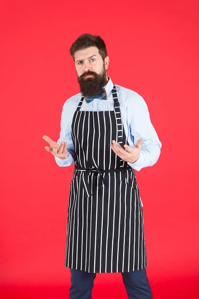 Comment puis-je vous aider ? Homme avec barbe cuisinier tablier hipster. Hipster chef cuisinier fond rouge. Un homme barbu cuisinier. Service et personnel du restaurant. Serveur bien entretenu au travail. Concept de café Hipster — Photo