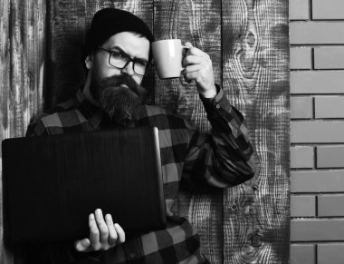 Sakallı, beyaz tenli hippi elinde bardakla dizüstü bilgisayar tutuyor.