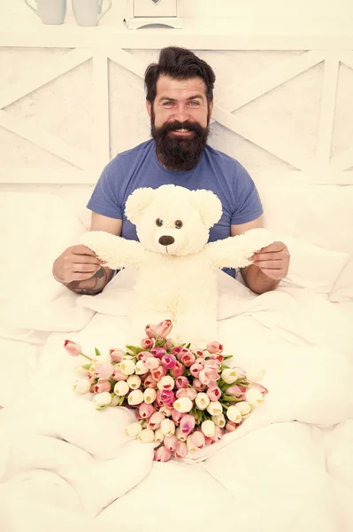 Birthday holiday. Make surprise concept. Gift for spouse. Teddy bear. Fresh flowers. Bearded hipster in bed. Valentines day gift. Man hold tulips bouquet relaxing in bed. Flowers delivery service