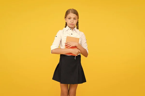What a sad news. Sad schoolgirl holding book on yellow background. Adorable little child in school uniform with sad emotion on face. Feeling sad and unhappy