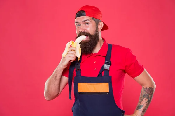 Morde banana. Um faz-tudo com fome. Faça um lanche saudável. Homem barbudo de uniforme e gorro comendo banana. Trabalhador construtor mecânico comer banana. Fruta fresca. Viver uma vida saudável. Conceito saboroso e gostoso — Fotografia de Stock