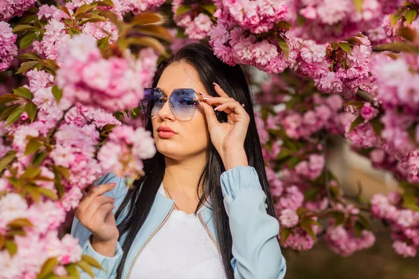 Bahar çiçeğinin açtığı süslü güneş gözlüklü kadın. Kiraz çiçekli kız. Bahar moda koleksiyonu. Sakura ağacı çiçek açıyor. Sakura ona yakışıyor. Muhteşem çiçek ve dişi güzellik. Aksesuar dükkanı. — Stok fotoğraf