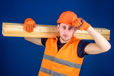 Koruyucu eldivenli adam miğfer takıyor, kafasındaki kaskı düzeltiyor, mavi arka plan. Odun işçisi konsepti. Marangoz, marangoz, marangoz, işçi, inşaatçı omuzunda tahta kiriş taşıyor.