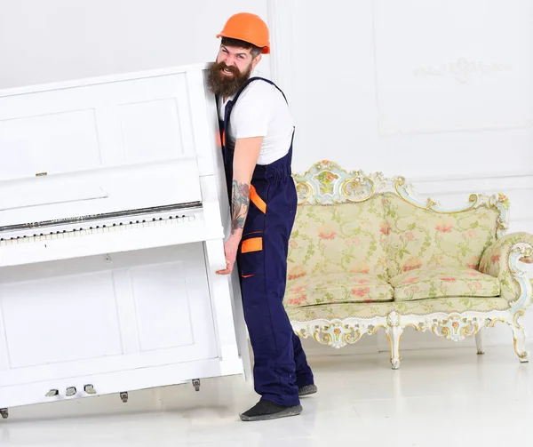 Loader moves piano instrument. Man with beard worker in helmet and overalls lifts up, efforts to move piano, white background. Courier delivers furniture, move out, relocation. Heavy loads concept.