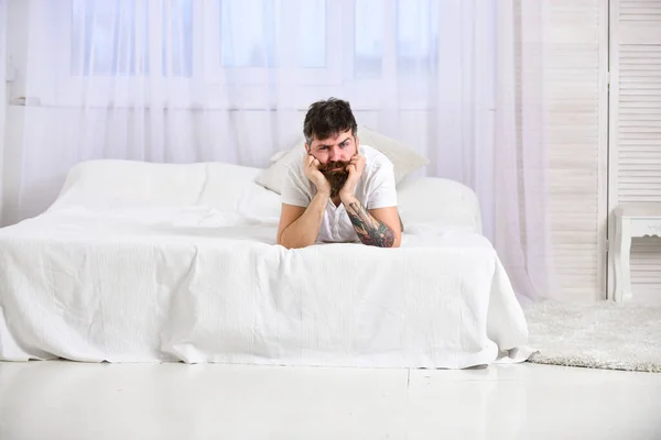 Guy on sleepy face laying on edge of bed on white sheets. Bedroom and mattress concept. Man laying on bed, white curtains on background. Macho with beard and mustache relaxing, having rest.