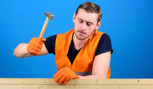 Carpenter, woodworker on busy face hammering nail into wooden board. Building and construction concept. Man, labourer, handyman in bright vest and protective gloves handcrafting, blue background