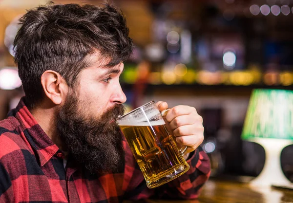 A fickó egy bárban tölti a szabadidejét, kitiltott háttérrel. Egy fáradt vagy nyugodt arcú férfi ül egy bárban vagy kocsmában a pult mellett. Hipster szakállas tartja üveg sör, italok, élvezze a friss íz. Pihenési koncepció — Stock Fotó