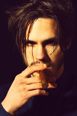 Ciddi sakallı bir adam siyah stüdyo arka planında puro içiyor.