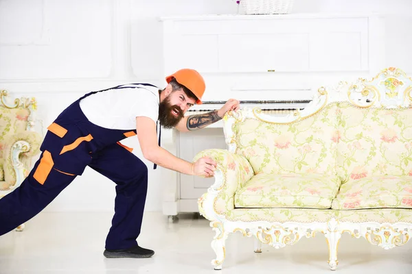 Delivery service concept. Courier delivers furniture in case of move out, relocation. Man with beard, worker in overalls and helmet pushes sofa, white background. Loader moves couch.