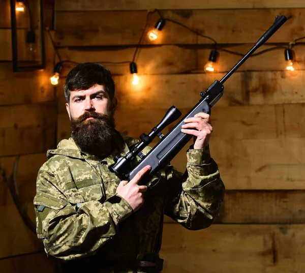 Man with beard wears camouflage clothing in wooden interior background. Hunter, brutal hipster with gun in his hand ready for hunting. Macho on strict face at gamekeepers house. Gamekeeper concept — Stock Photo, Image