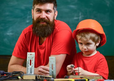 Baba, sakallı bir ebeveyn ve küçük oğlu sınıfta, arka planda kara tahta var. Koruyucu miğferli çocuk elleriyle tamir ediyor, babasıyla el işi yapıyor. Baba tamirci konsepti.