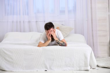 Uykulu esneyen suratlı adam beyaz çarşafların üzerinde yatıyordu. Yatak odası konsepti. Sakallı ve bıyıklı maço, dinleniyor. Yatakta yatan bir adam, arka planda beyaz perdeler.