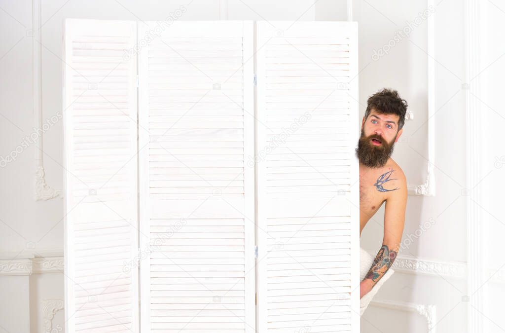 Man, lover in white interior caught naked. Lovers and cheating concept. Man with beard and mustache hiding behind folding screen. Hipster naked on shocked face unexpectedly detected in bedroom