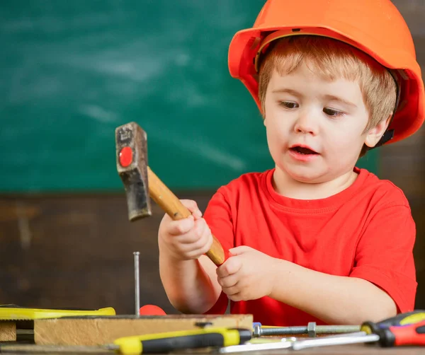 Kind mit Helm spielt als Bauarbeiter oder Reparateur, repariert oder bastelt. Handwerkliches Konzept. Kleinkind auf viel beschäftigtem Gesicht spielt zu Hause in der Werkstatt mit Hammerwerkzeug. Junge spielt als Heimwerker — Stockfoto