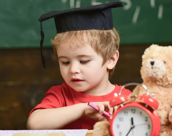 Elementary education concept. First former with toy on desk, close up. Pupil in mortarboard, chalkboard on background. Kid studies near alarm clock and teddy bear. Boy smart, clever, likes studying