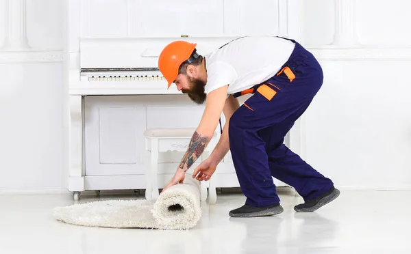 Relocating concept. Courier delivers furniture in case of move out, relocation. Loader wrappes carpet into roll. Man with beard, worker in overalls and helmet rolling carpet, white background