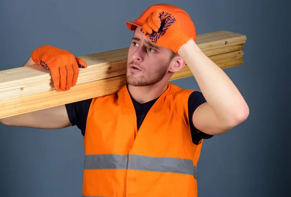 Man in protective gloves holds visor of protective hard hat, grey background. Safety and protection concept. Carpenter, woodworker, strong builder on serious face carries wooden beam on shoulder. — Stock Photo, Image