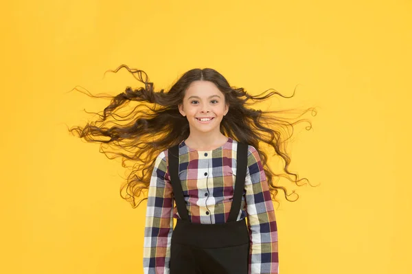 Pura belleza para tu cabello. Niño feliz con el pelo largo ondulado. Niño pequeño con el pelo volador de fondo amarillo. Mirada escolar de niña pequeña. Peluquería. Volver a la escuela moda uniforme —  Fotos de Stock