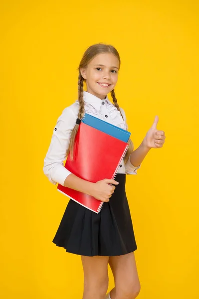 다시 학교로. 제복을 입은 작은 소녀가 엄지 손가락을 보여 줍니다. 수업이 끝 날 때마다 문서를 가지고 다닌다. 집에서 공부 한다. 학교의 패션 스타일. 노란색 배경을 가진 똑똑 한 학생처럼 보이죠. 현대 교육 개념 — 스톡 사진