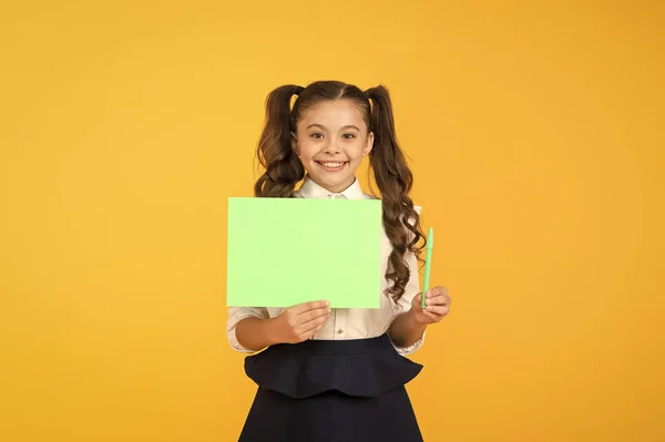 Skaffa en Skriv penna. Liten flicka som håller tomma pappersblad för att skriva uppgift på gul bakgrund. Små barn Visa grön affisch för att skriva information. Tomt Visa kort för att skriva, kopiera utrymme — Stockfoto