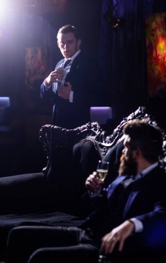 Smokinli seksi adam elinde bir bardak şampanyayla bir gece kulübünün karanlığında oturuyor..