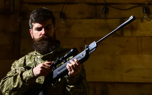 Man with beard wears camouflage clothing, wooden interior background. Huntsman concept. Macho on strict face at gamekeepers house. Hunter, brutal hipster with gun prepare rifle for hunting, copy space — Stock Photo, Image