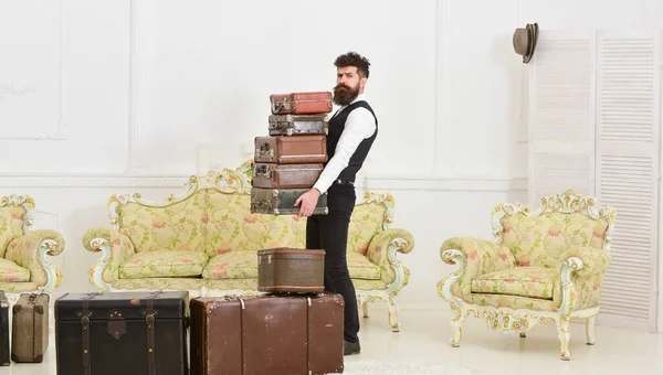 Macho elegante en la cara estricta lleva montón de maletas vintage. Butler y concepto de servicio. Hombre con barba y bigote con traje clásico entrega equipaje, fondo interior blanco de lujo . — Foto de Stock