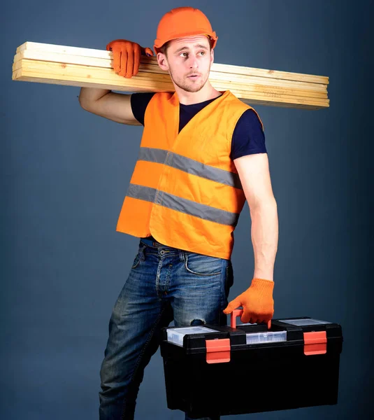 Concept de menuiserie. Homme au casque, casque rigide tient boîte à outils et poutres en bois, fond gris. Menuisier, menuisier, ouvrier, constructeur sur le visage calme porte des poutres en bois sur l'épaule . — Photo