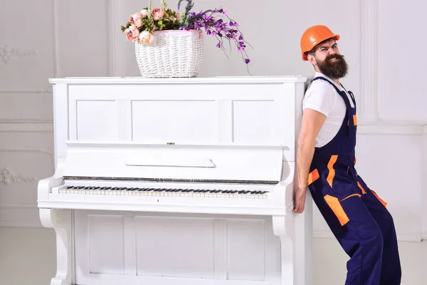 El mensajero entrega muebles, se muda, se traslada. Hombre con barba trabajador en casco y overoles empuja, poner esfuerzos para mover el piano, fondo blanco. El cargador mueve el instrumento de piano. concepto de cargas pesadas . — Foto de Stock