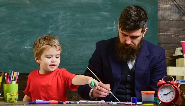 ひげを生やした教師、父は教室で描くために小さな息子を教える、背景に黒板。才能あるアーティストは息子と一緒に時間を過ごす。忙しい顔の絵に子供や教師、描画します。美術の授業のコンセプト. — ストック写真