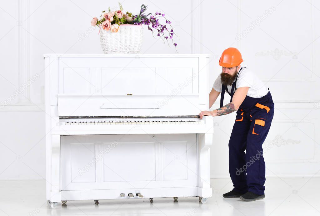 Busy worker in orange protective helmet and blue overall moving grand piano isolated on white background. White piano with flower vase on top in empty room