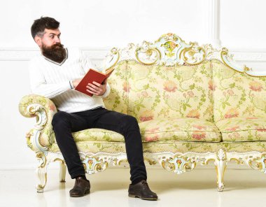 En çok satan skandallar konsepti. Adam şaşkınlıkla kitap okuyor. Şaşırmış yüz okuma kitabı üzerine maço. Sakallı ve bıyıklı bir adam barok tarzı koltukta oturuyor, kitap tutuyor, arka planda beyaz duvar var..