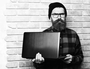 Sakallı, beyaz tenli hippi bej tuğlalı stüdyo arka planında dizüstü bilgisayar tutuyor.