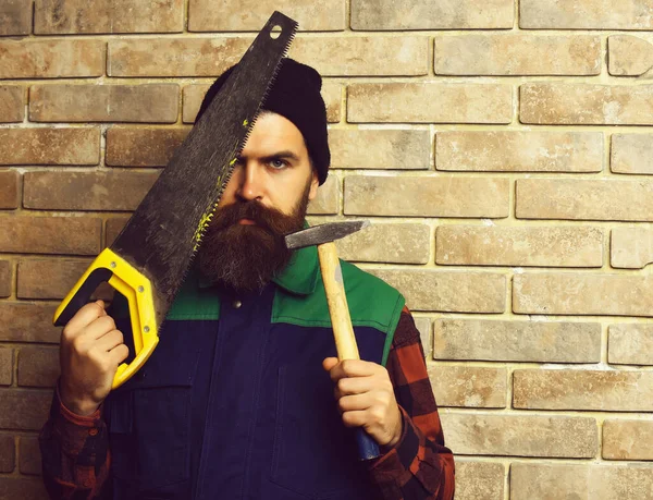 Bärtiger Arbeiter hält Säge und Hammer mit ernstem Gesicht — Stockfoto