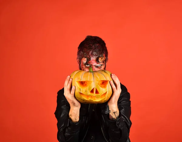 Man wearing scary makeup holds pumpkin on red background.