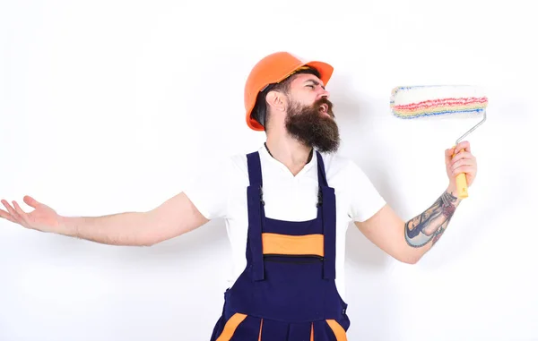 Opknappen. Plezier op het werk. Man met baard in oranje beschermhelm met verfroller als microfoon. Jonge bouwer die zingt alsof hij een rockster is — Stockfoto