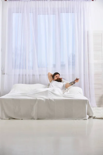 Egy inges férfi fekszik az ágyon, tévét néz, fehér függönyök a háttérben. Egy fickó komoly arccal távirányítóval kapcsolja a csatornát. Macho szakállal tartja az irányítót a kezében. Távirányítási koncepció. — Stock Fotó