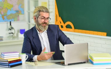 Resmi başvuru çevrimiçi. Okul ödevi. Okula dönelim. Bilgi toplama günü. Erkek işçi okulu öğretmeni sınıfta oturur. Yakışıklı hippi, olgun, akıllı eğitimci. Dizüstü bilgisayar ve internet kullan