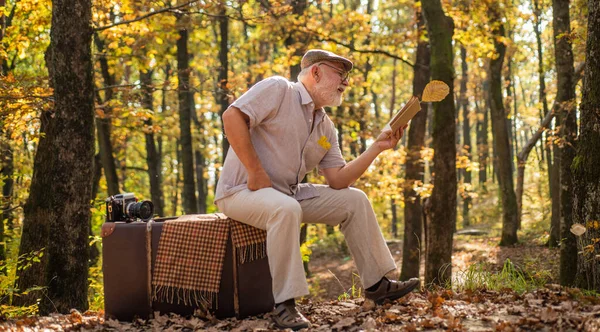 literature outside of library. getting knowledge and information from nature. Old man read sitting on retro suitcase. autumn trip. Having leisure to read more. Retired man read book in autumn nature