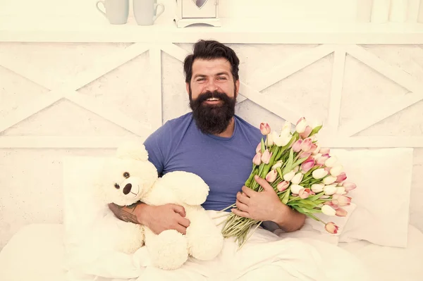 Birthday holiday. Fresh flowers. Bearded hipster in bed. Valentines day gift. Man hold bouquet relaxing in bed. Flowers delivery service. Make surprise concept. Gift for spouse. Cute teddy bear