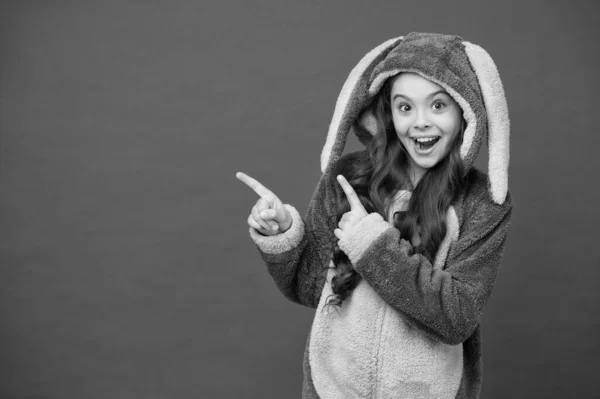 Take a look. Happy girl pointing index fingers at something. Small child in bunny pajamas pointing red background. Pointing gesture. Pointing and promoting. For sale, copy space — Stock Photo, Image