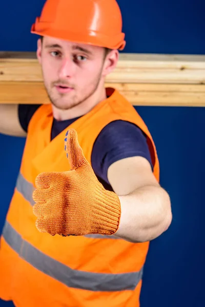 Carpintero, carpintero, constructor en la cara estricta lleva viga de madera en el hombro. Concepto de materiales de madera. Hombre en casco y guante protector muestra gesto pulgar hacia arriba, fondo azul, enfoque selectivo — Foto de Stock